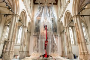 Een man op een rode hoogwerker voor het orgel in De Nieuwe Kerk die is ingepakt met lichtdoorzichtig plastic ter bescherming tegen het stof dat vrijkomt bij werkzaamheden.